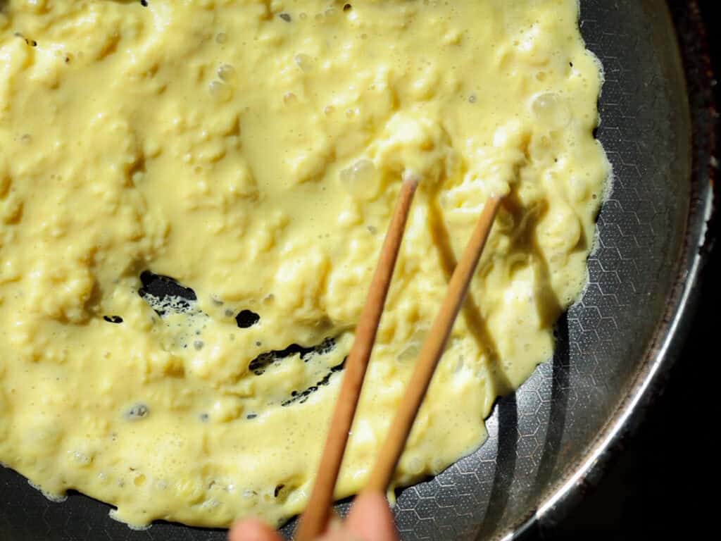 Close-up of scrambled vegan eggs being stirred with chopsticks in a non-stick frying pan. The eggs are partially cooked, with a creamy texture and pale yellow color, indicating they are still in the process of being prepared.